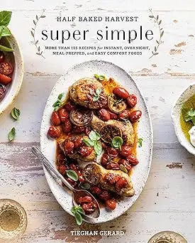 A white cookbook half baked harvest super simple on a rustic wooden table with a platter of roasted chicken garnish