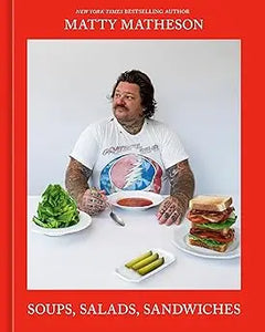 A man with tattoos wearing a white t-shirt with a colorful graphic design seated at a table with salads and sandwiches in matty matheson